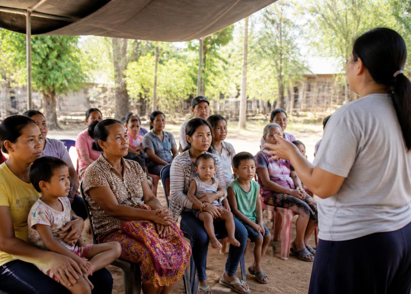 Families seated under a shaded canopy listening to a community health facilitator